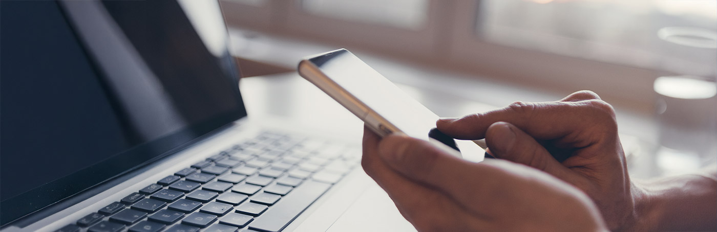 Image of a person scrolling news on their phone, with their laptop on in the background